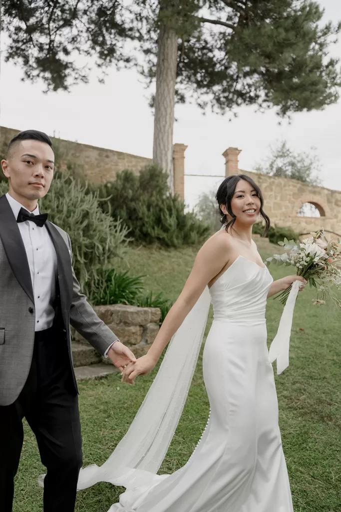 Bride and groom stroll hand in hand across the courtyard at Borgo Sant Ambrogio, with stone walls and lush greenery.
