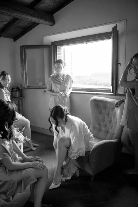 Bride surrounded by bridesmaids while putting on her shoes in a sunlit room at Borgo Sant Ambrogio.