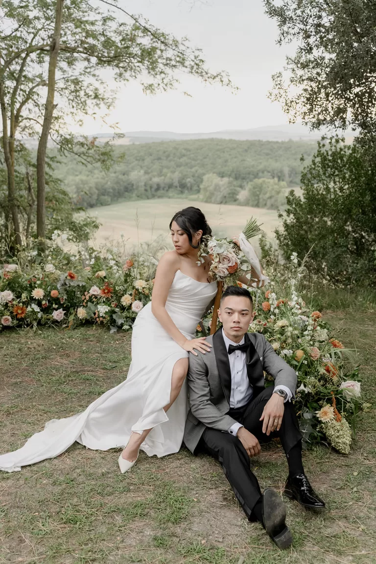 Bride and groom seated in a floral meadow overlooking the forested hills of Borgo Sant'Ambrogio.