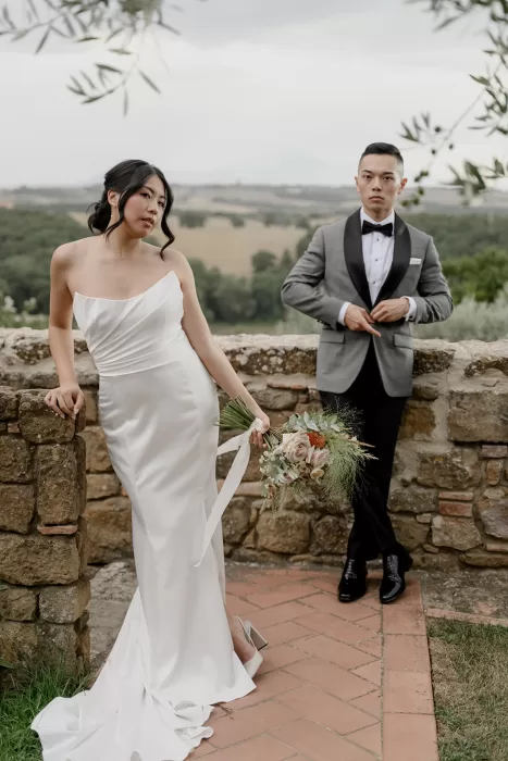 Bride leans against a stone wall holding her bouquet while the groom adjusts his jacket at Borgo Sant'Ambrogio.