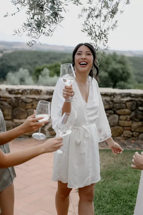 Bride in white robe raises her glass in a joyful toast under olive branches at Borgo Sant'Ambrogio.