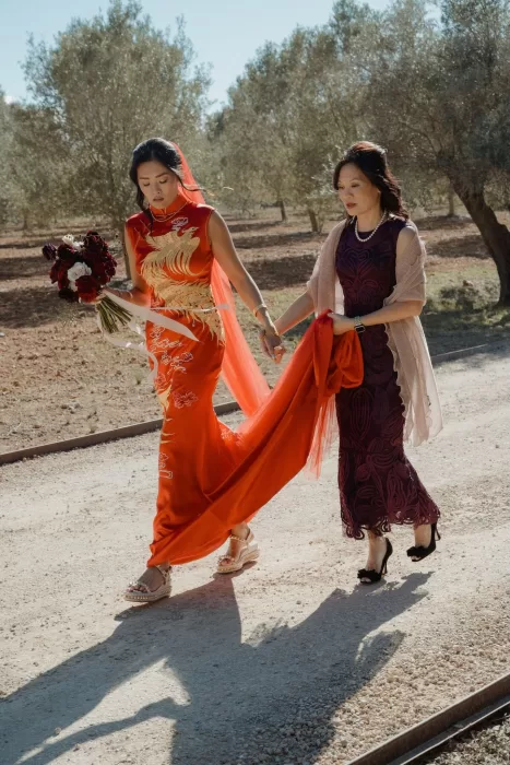 Bride and her mother walk hand in hand in elegant cheongsams at Ca Na Xica, under a clear blue sky.