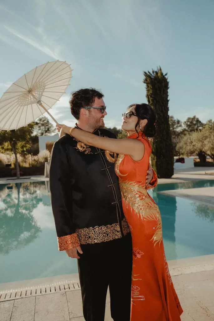 Bride and groom sharing a joyful moment by the pool at Ca Na Xica, with a parasol and traditional outfits.