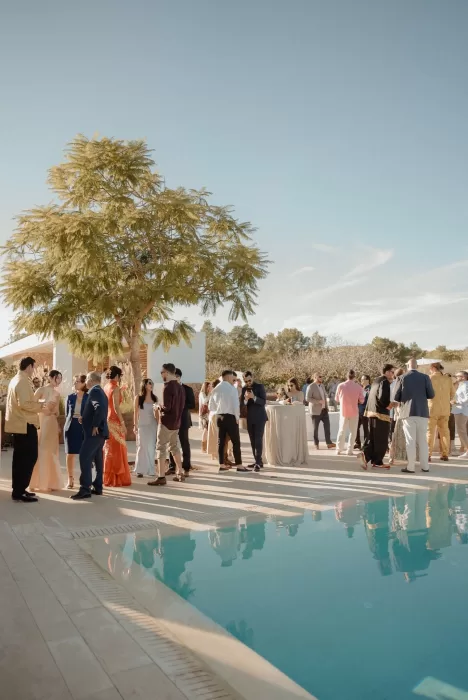 Wedding guests mingling by the pool during cocktail hour at Ca Na Xica, surrounded by warm light and trees.