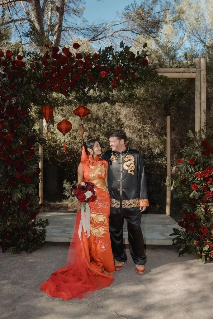 Couple standing beneath a floral arch with red lanterns at Ca Na Xica, surrounded by forest greenery.