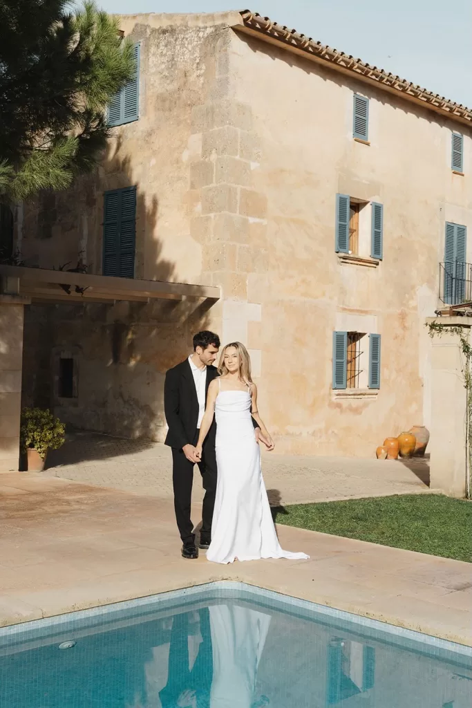 Bride and groom standing hand in hand by the pool with rustic stone walls and Mediterranean details at Finca Son Sampol.