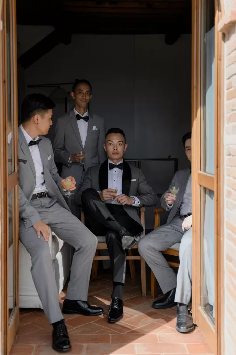 Groomsmen relax indoors at Borgo Sant'Ambrogio, dressed in grey suits and sipping cocktails before the ceremony.