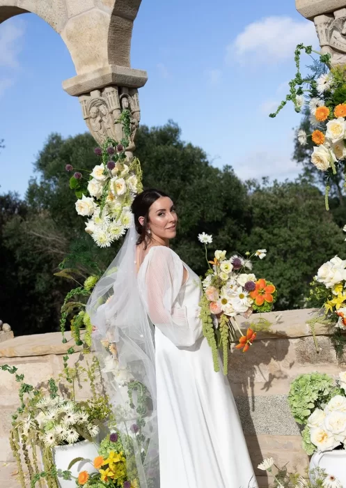The bride smiles over her shoulder, holding a whimsical bouquet beneath Mas Torroella’s arches.