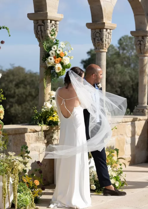 Back view of the bride’s open-back gown and long veil flowing in the breeze during the ceremony at Mas Torroella.