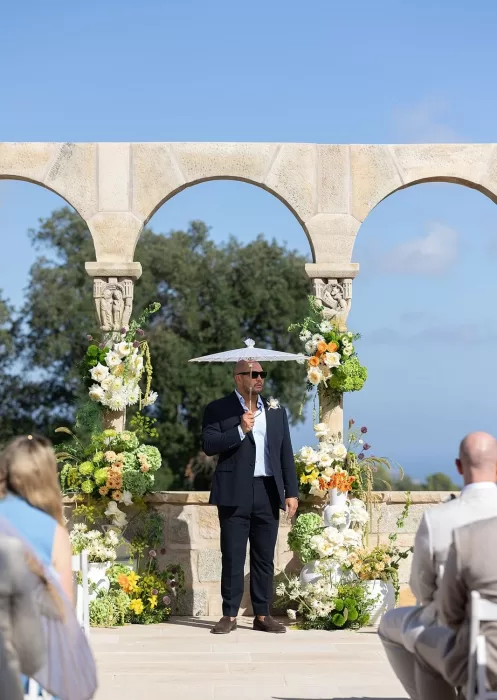 A guest stands beneath Mas Torroella’s arched stone ceremony setup, framed by lush floral arrangements and clear skies.