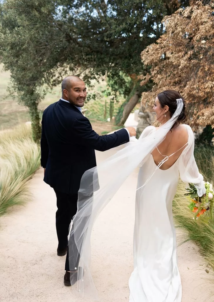The groom glances back with a smile as the couple walk hand-in-hand along a garden path at Mas Torroella.