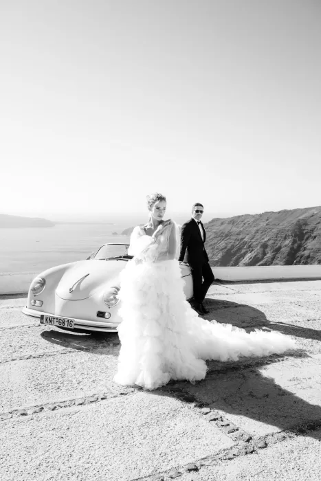 Editorial black and white portrait of a bride in textured gown and groom by a vintage car at Rocabella Santorini.