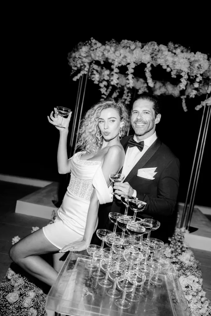 Playful black and white photo of bride and groom toasting beside a champagne tower during their Rocabella Santorini celebration.