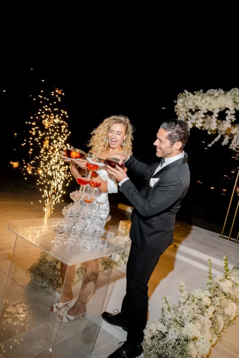 Bride and groom joyfully pour champagne into a tower under night sky at Rocabella Santorini celebration.