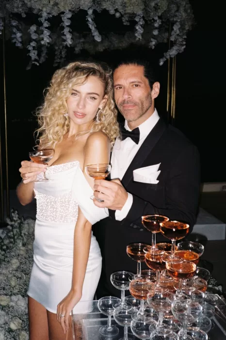Bride in pearl-embellished mini dress and groom pose with champagne glasses during a chic reception at Rocabella Santorini.