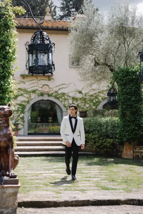 Groom walking toward the camera in front of the ivy-covered villa at Tenuta Corbinaia on a sunny day.