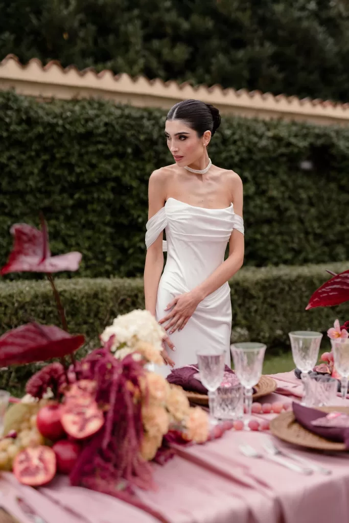 Bride in off-shoulder gown surrounded by floral details and pomegranates at an outdoor reception table at Villa Buttafava.