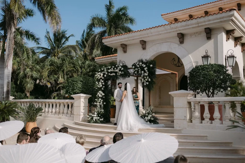 Romantic outdoor ceremony at Villa La Joya beneath a lush floral arch and palm trees.