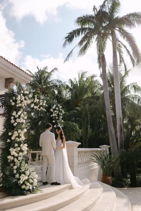 Couple exchanging vows beneath a white floral arch at Villa La Joya surrounded by palms.