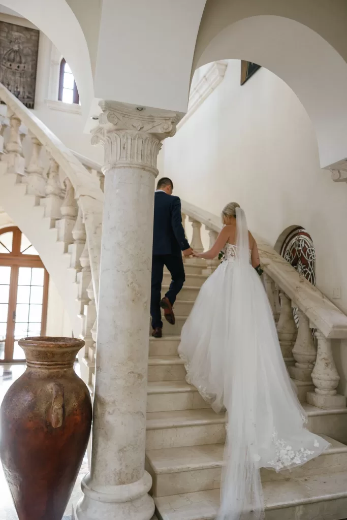 Bride and groom walk hand in hand up the grand stone staircase inside Villa La Joya.