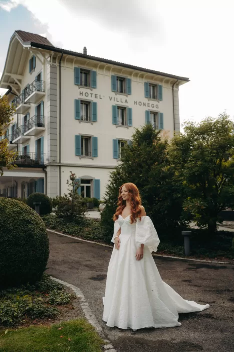 Bride in elegant white gown stands in front of Hotel Villa Honegg, framed by manicured gardens and soft light.