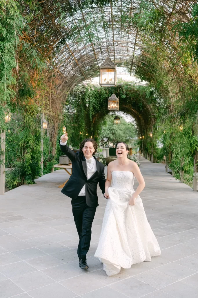 Newlyweds walking through lush garden archway filled with lanterns and vines at Luna Escondida.