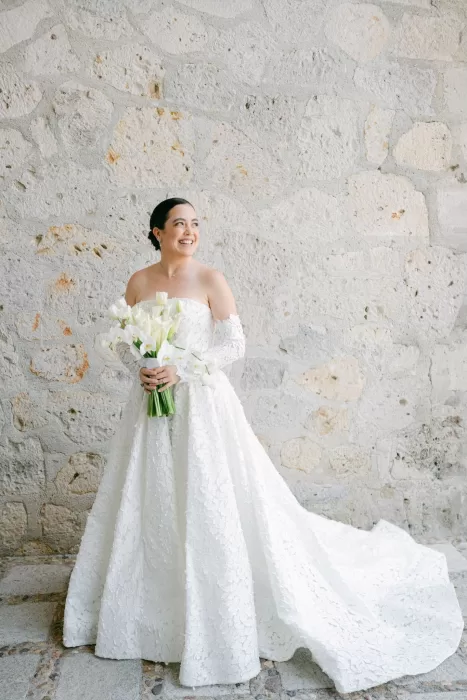 Smiling bride in a floral lace gown holding a tulip and orchid bouquet against a stone wall at Luna Escondida