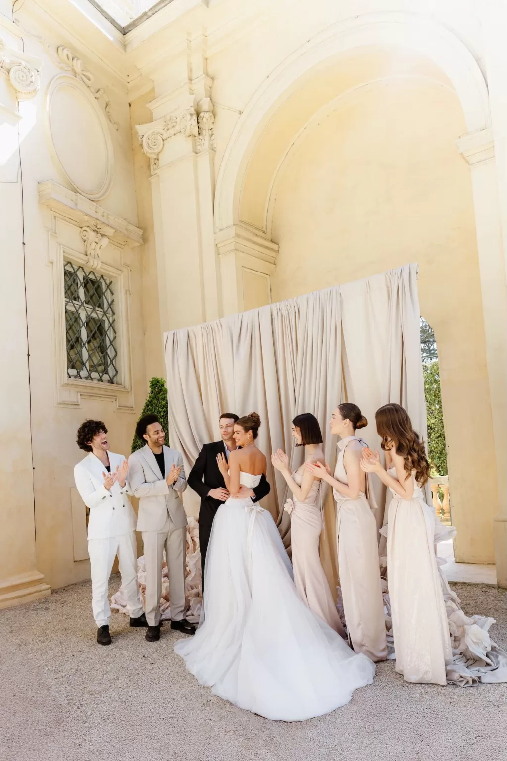 Bride and groom embrace in front of their bridal party inside a classical Italian villa, captured with warmth and joy.