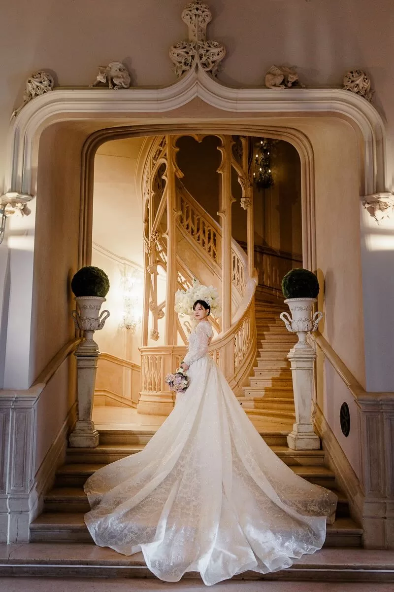 A bride stands poised at the base of a sweeping staircase in a fairytale setting, captured by Wim Vanhengel.