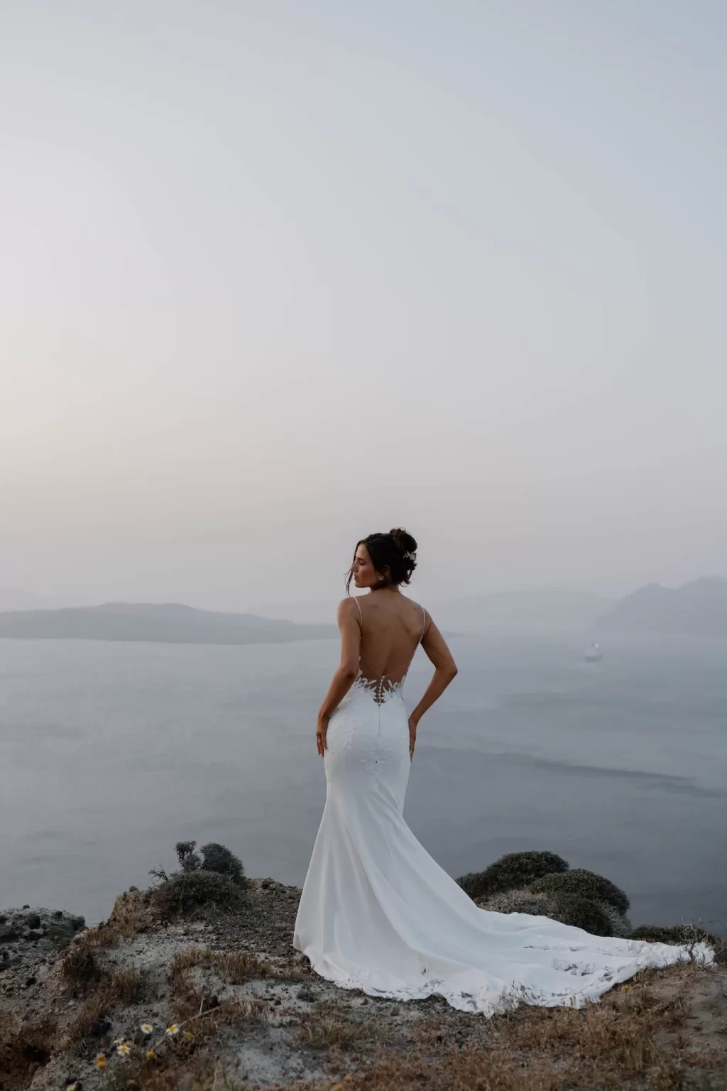 Bride stands confidently on a clifftop in Santorini with her train flowing behind her, gazing into the sunset, by Bahr Studios.