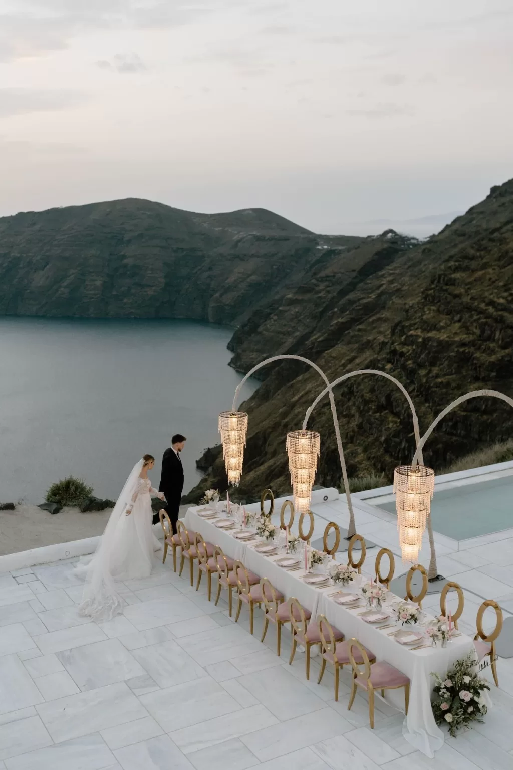 Bride and groom arrive at an elegant candlelit reception table perched above the sea in Santorini, captured by Bahr Studios.