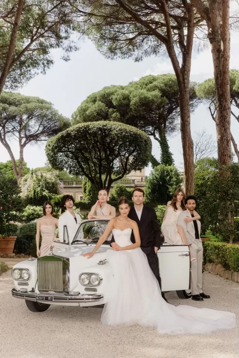 The wedding party poses with a vintage white car under the pines at Villa Aurelia’s garden entrance.