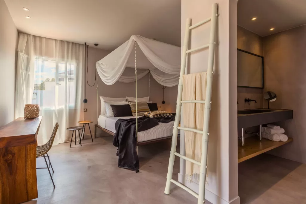 Canopy bedroom at Minos Beach Art Hotel with flowing white drapes, warm wood accents, and an ensuite bathroom.