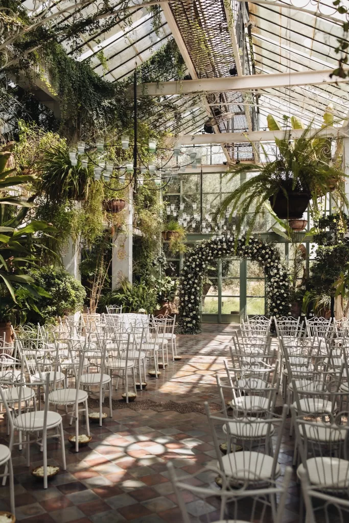 eremony setup inside the glasshouse at Beldi Country Club, with white chairs, chandeliers, and a floral arch under natural light.