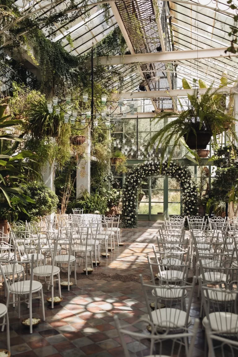 eremony setup inside the glasshouse at Beldi Country Club, with white chairs, chandeliers, and a floral arch under natural light.
