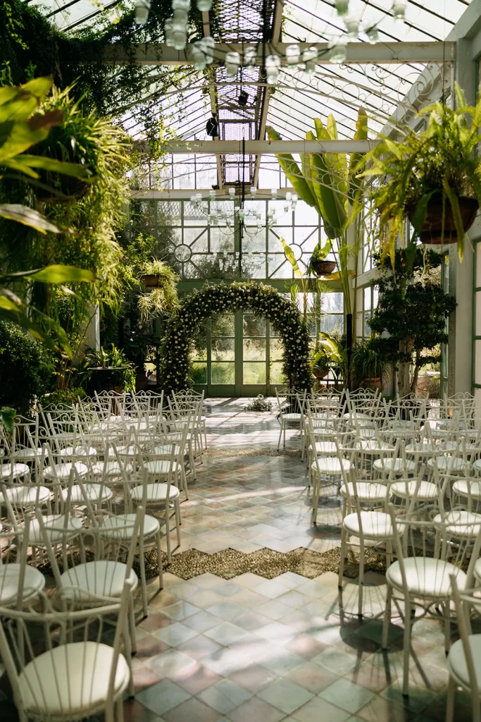 Sunlit ceremony setup inside the glasshouse at Beldi Country Club, surrounded by lush greenery and white chairs.