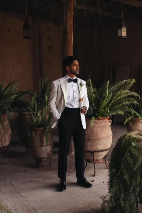 Groom in white tuxedo jacket holding champagne glass among terracotta pots at Beldi Country Club.