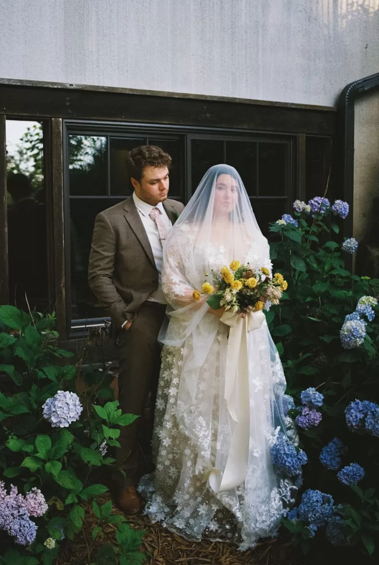 Romantic portrait of bride and groom by a window surrounded by blue hydrangeas at House Boheme.