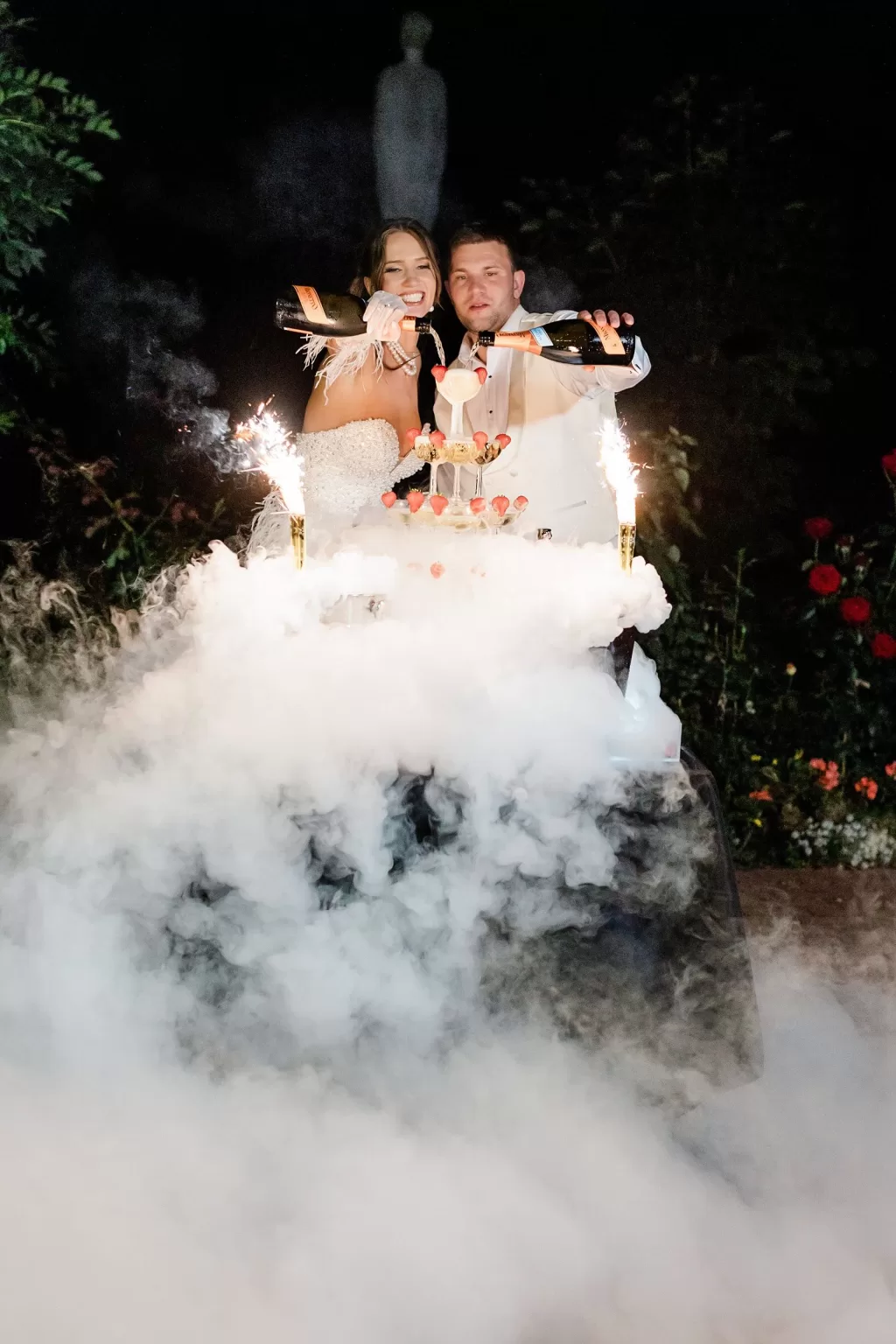 Newlyweds pouring champagne over a tower with sparklers and smoke effects during night celebration, photographed by Jurgita Lukos.