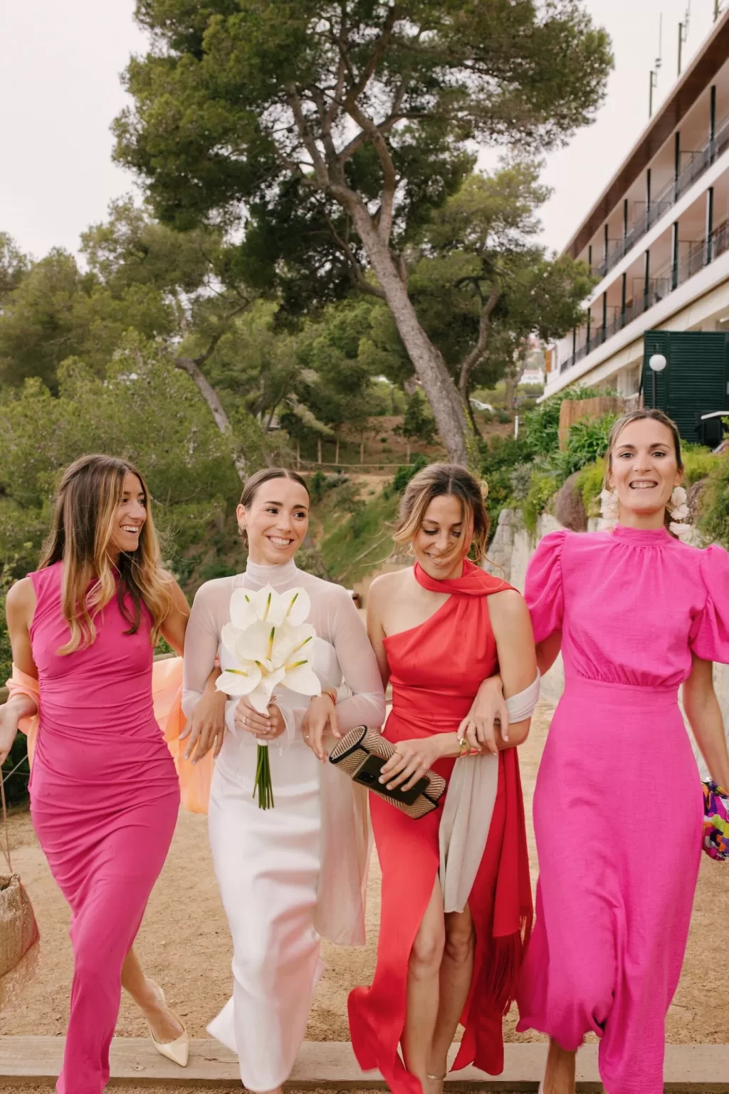 Smiling bride walking arm in arm with bridesmaids in bold dresses at an outdoor wedding, photographed by Lucky Elevens.