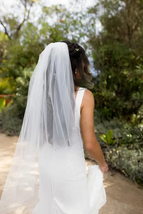 Bride in lace gown with long veil walking through the lush pathways of Marimurtra Botanical Garden.