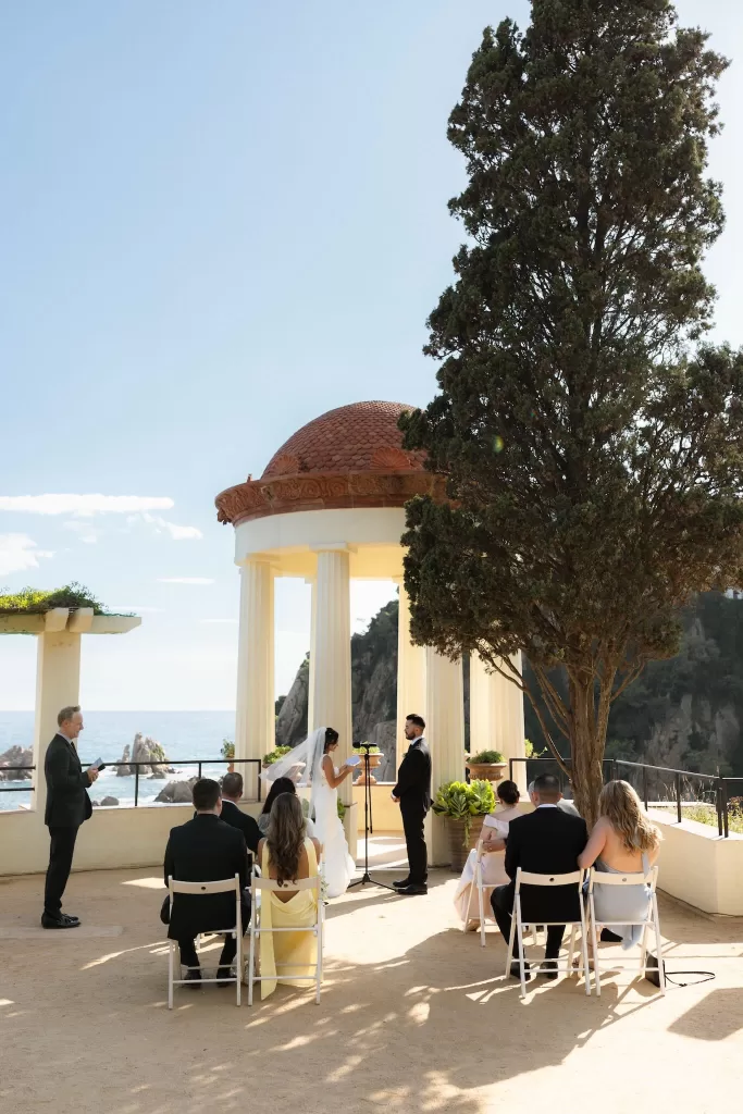 Wedding ceremony with bride and groom exchanging vows by the pavilion at Marimurtra Botanical Garden, overlooking the sea.