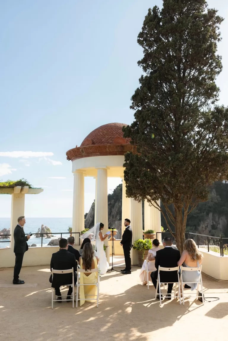 Wedding ceremony with bride and groom exchanging vows by the pavilion at Marimurtra Botanical Garden, overlooking the sea.