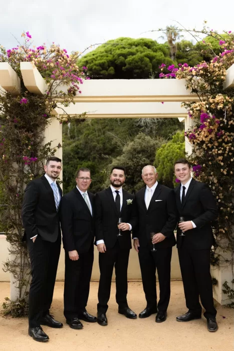 Groom with groomsmen dressed in black suits standing under floral arch at Marimurtra Botanical Garden.