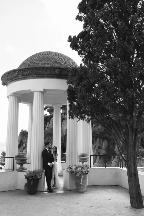 Bride and groom standing by the grand pavilion of Marimurtra Botanical Garden with a tree in the foreground.