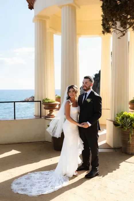 Couple standing together by the seaside pavilion at Marimurtra Botanical Garden, with ocean views behind.
