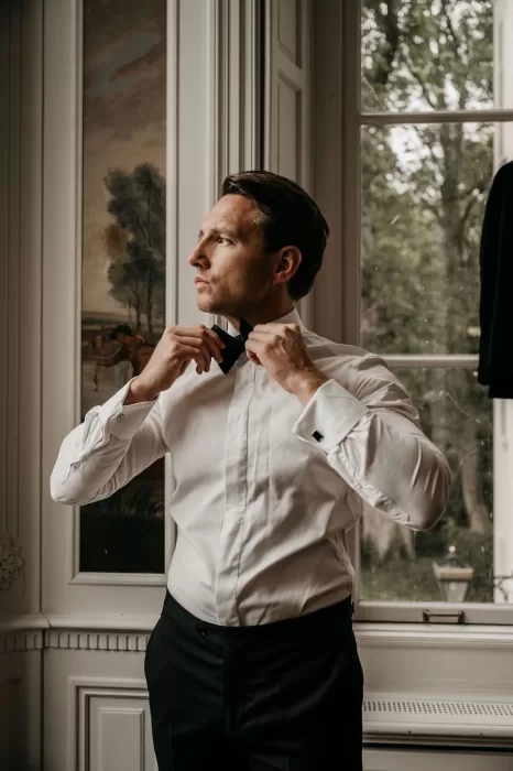 Groom adjusting his bow tie by the window during preparations at Parc Broekhuizen.