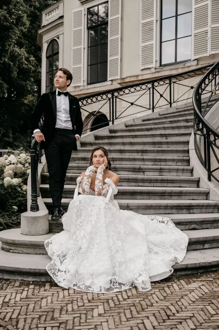 Bride seated in her gown on the staircase of Parc Broekhuizen with groom standing beside her.