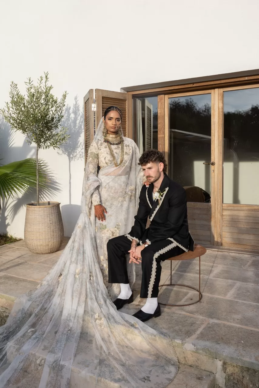 Bride in embroidered saree and groom in tailored suit pose together outdoors, by Sus Pons.