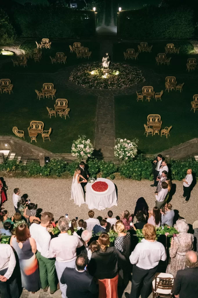 Bride and groom cutting their wedding cake outdoors at night surrounded by guests in the gardens of Villa Bossi.